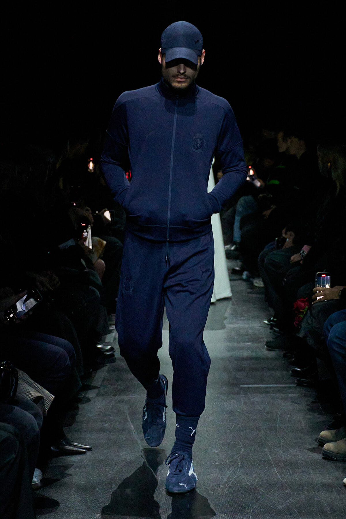 Male model wearing a navy tracksuit and cap walks a dim fashion runway, hands in pockets, focused forward.