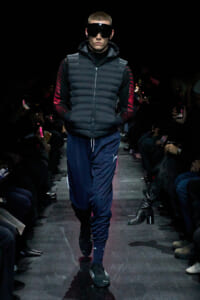 Male model on a runway wearing a black padded vest with a hood, oversized black goggles, navy track pants, and black sneakers; audience seated along the catwalk.