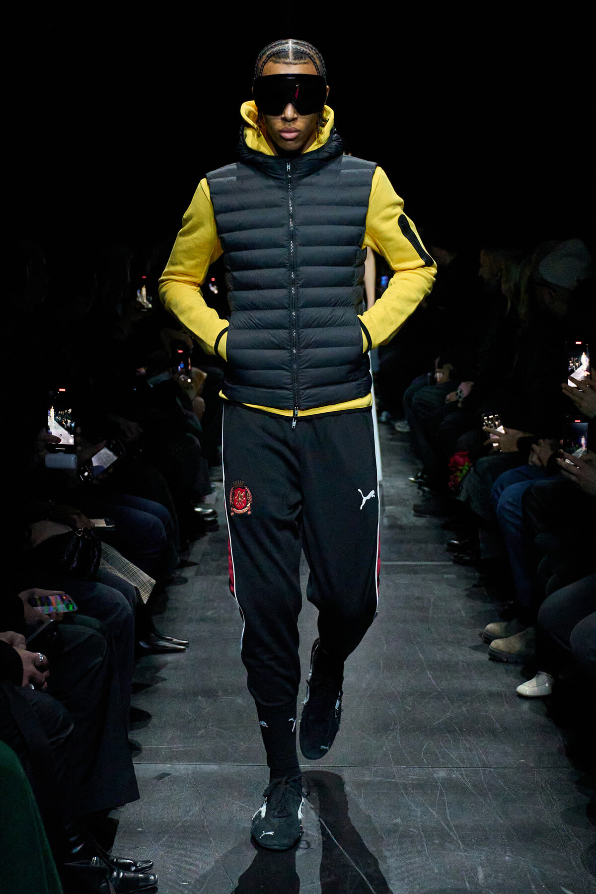 Model walks the runway in a yellow hoodie under a black puffer vest with braided hair and oversized sunglasses, audience in darkness.