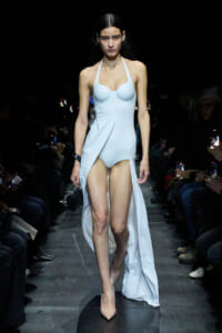 Model walks a fashion show wearing a pale blue halter-neck gown with thigh-high slits, beige heels, and a small black clutch in one hand.