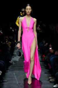 Model walking a fashion runway in a bright pink halter dress with a high slit, carrying a small black handbag and wearing nude heels.