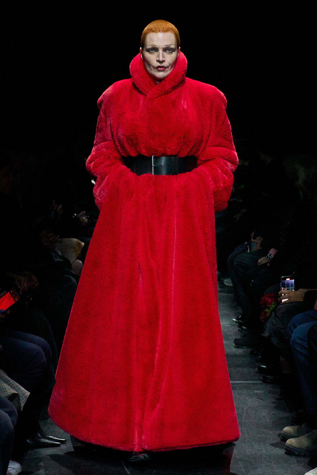 Model walks a runway in a bright red oversized fur coat cinched with a wide black belt against a dark background.