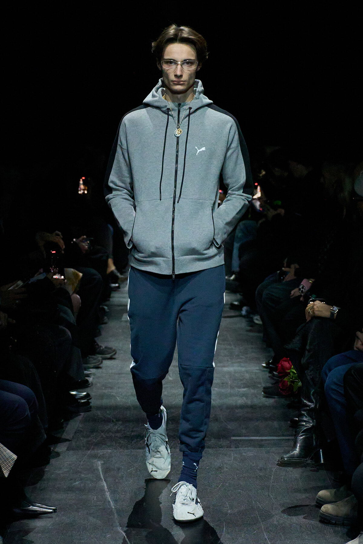 Male model on a dim runway wearing a gray zip hoodie with black panels, navy track pants, and white sneakers.