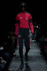 Male model walking a fashion runway in a red and black Puma tracksuit with crest emblem, sunglasses, and gloves on a dark runway.