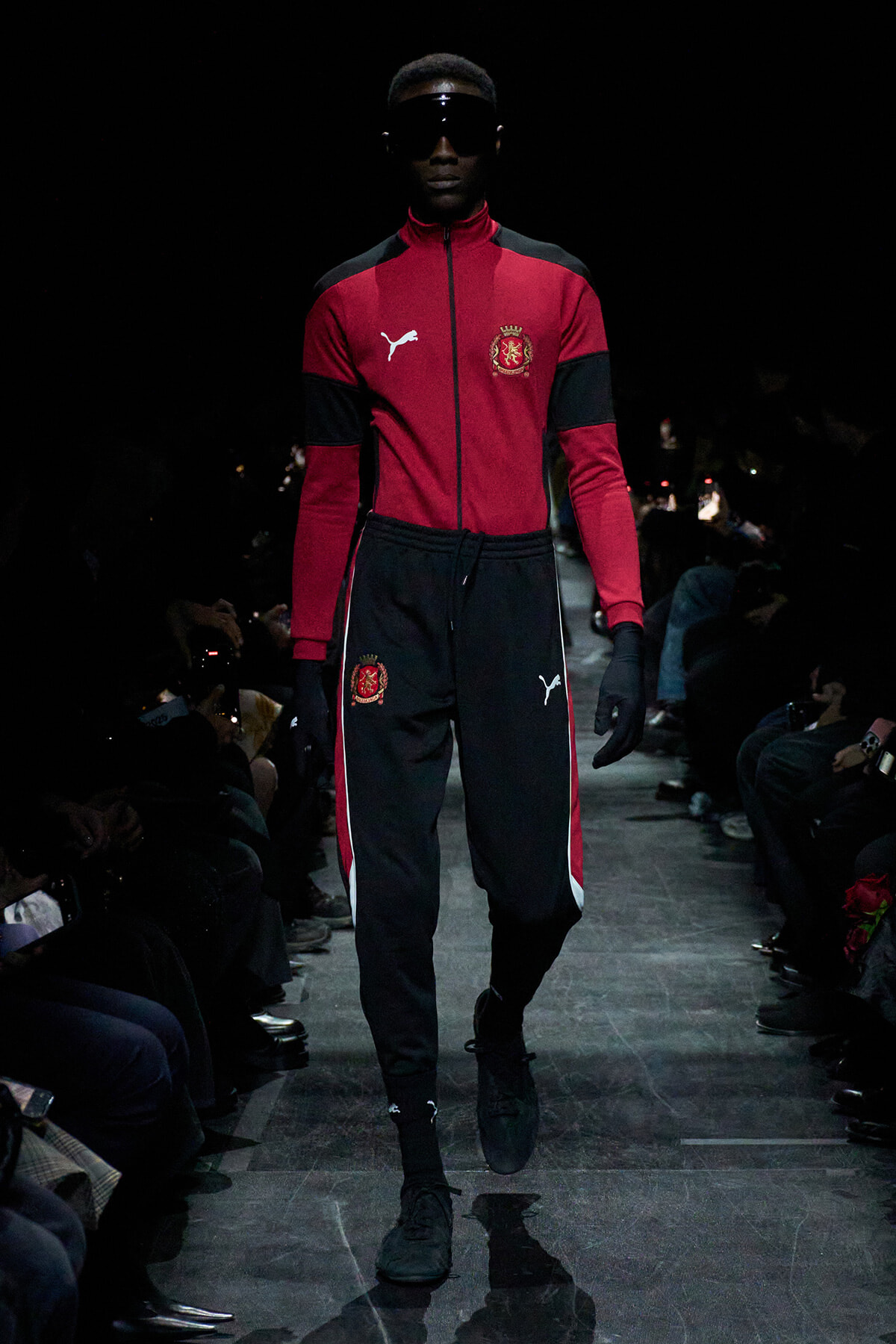 Male model walking a fashion runway in a red and black Puma tracksuit with crest emblem, sunglasses, and gloves on a dark runway.