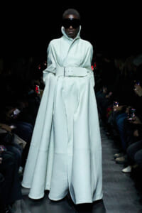 Model on a fashion runway wearing a long pale blue coat with a wide belt and high collar, paired with oversized black visor sunglasses, dim audience backdrop on both sides.