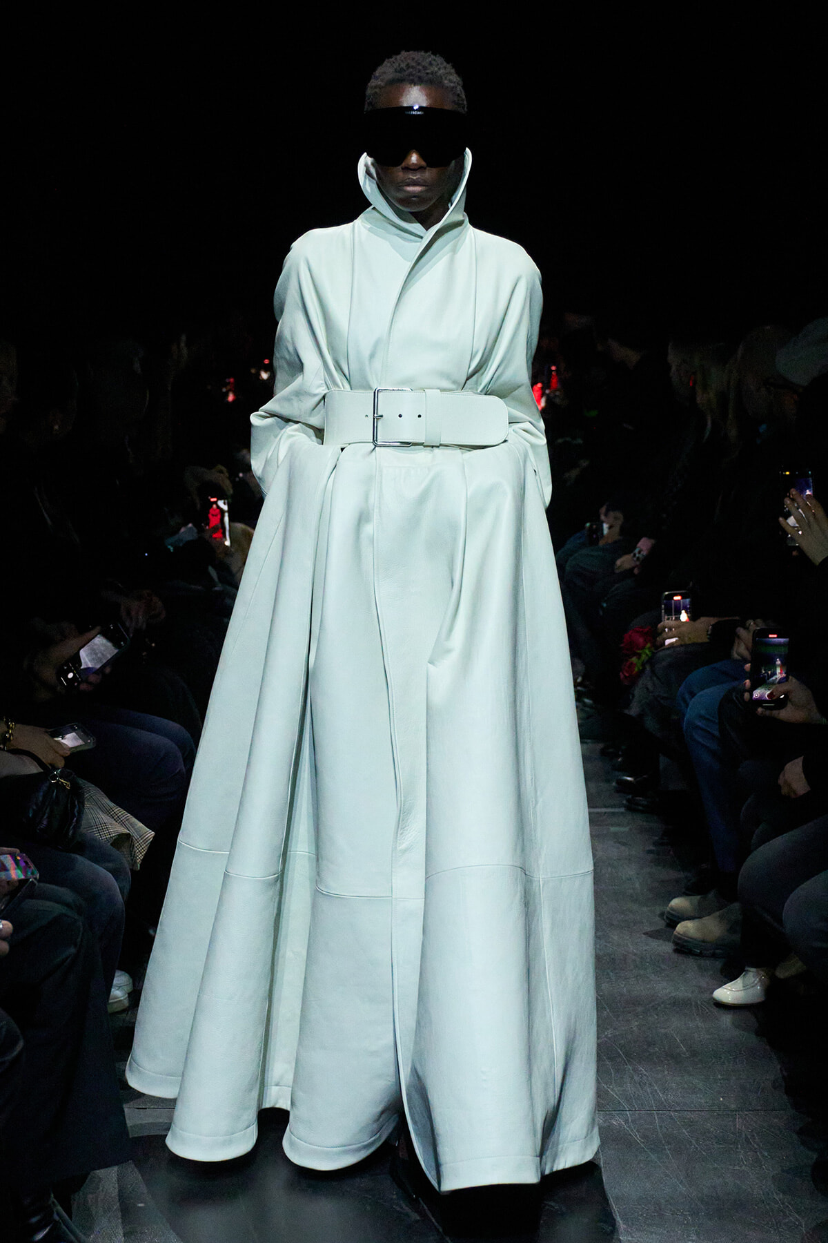 Model on a fashion runway wearing a long pale blue coat with a wide belt and high collar, paired with oversized black visor sunglasses, dim audience backdrop on both sides.