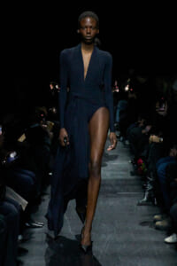 Model walks the runway in a navy draped wrap dress with a deep V-neck and high side slit, surrounded by an audience in a dark venue.