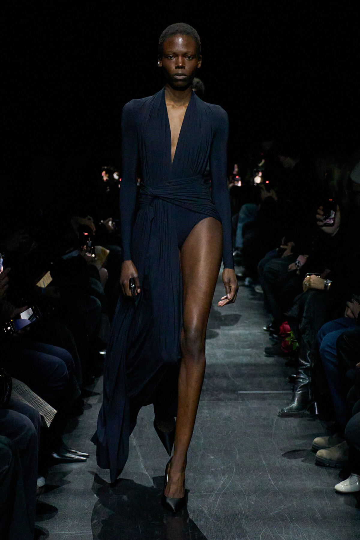 Model walks the runway in a navy draped wrap dress with a deep V-neck and high side slit, surrounded by an audience in a dark venue.