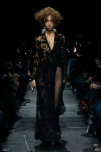 Model walks a fashion show wearing a sheer black lace gown with a deep V, sequins, and a high slit; tousled short hair and a necklace visible in the spotlight.