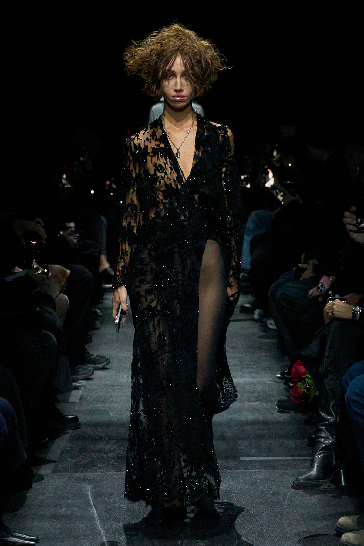 Model walks a fashion show wearing a sheer black lace gown with a deep V, sequins, and a high slit; tousled short hair and a necklace visible in the spotlight.