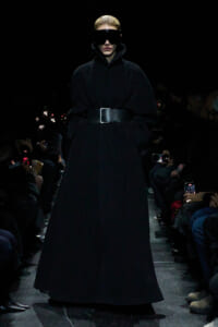Runway model in a floor-length black coat cinched with a wide belt and oversized black visor sunglasses.