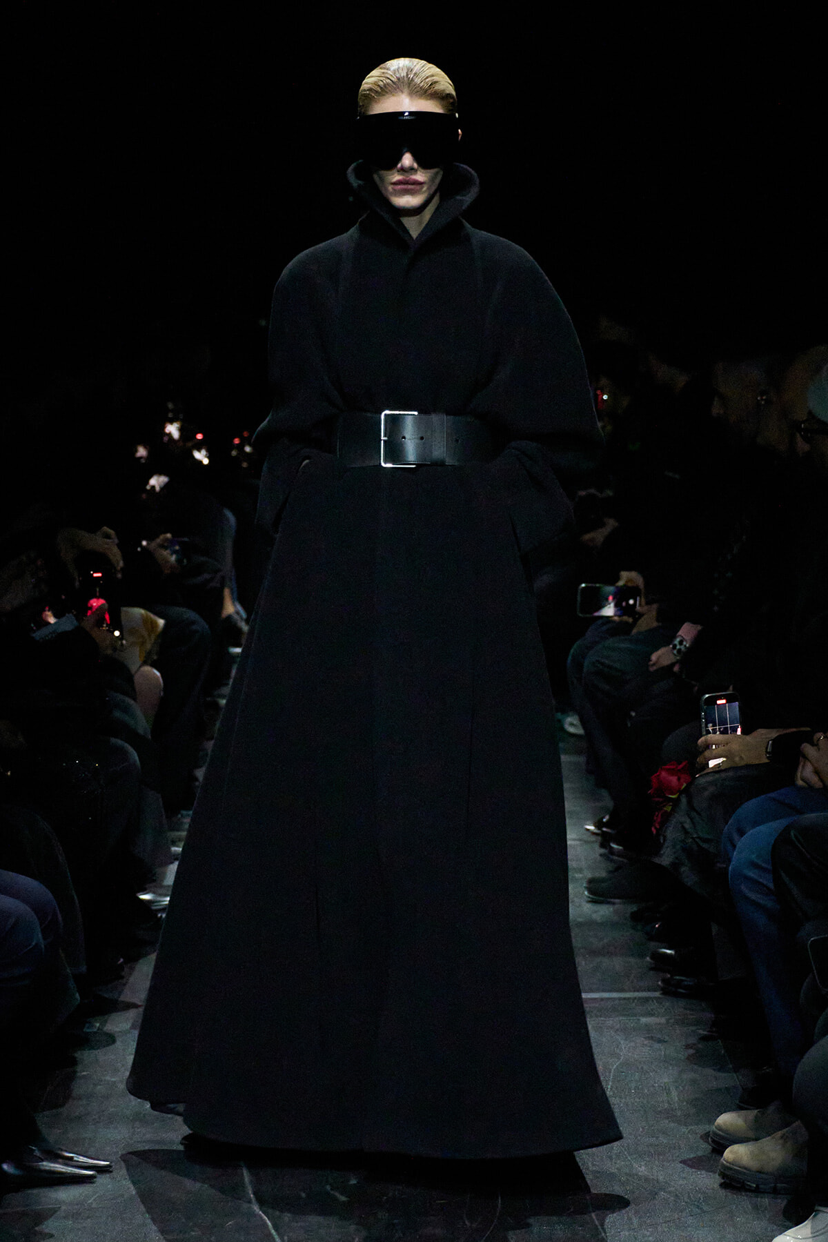 Runway model in a floor-length black coat cinched with a wide belt and oversized black visor sunglasses.