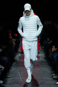 Model walking a fashion runway in an all-white padded jacket with hood, white cap, matching joggers and sneakers; dark audience sides.