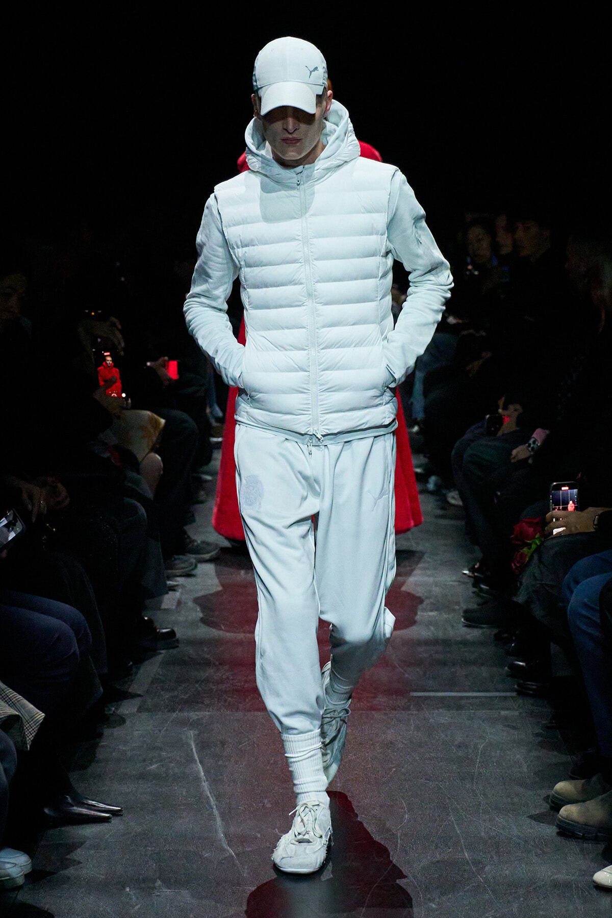 Model walking a fashion runway in an all-white padded jacket with hood, white cap, matching joggers and sneakers; dark audience sides.