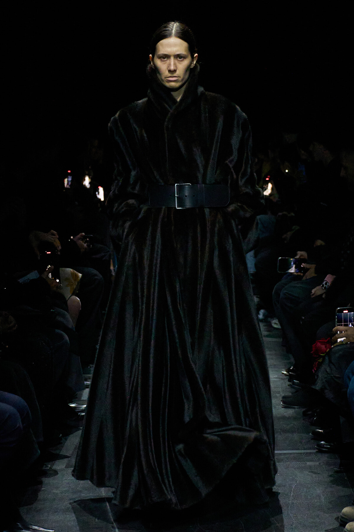 Model walking the runway in a long black fur coat with a wide belt, audience filming from the sides.