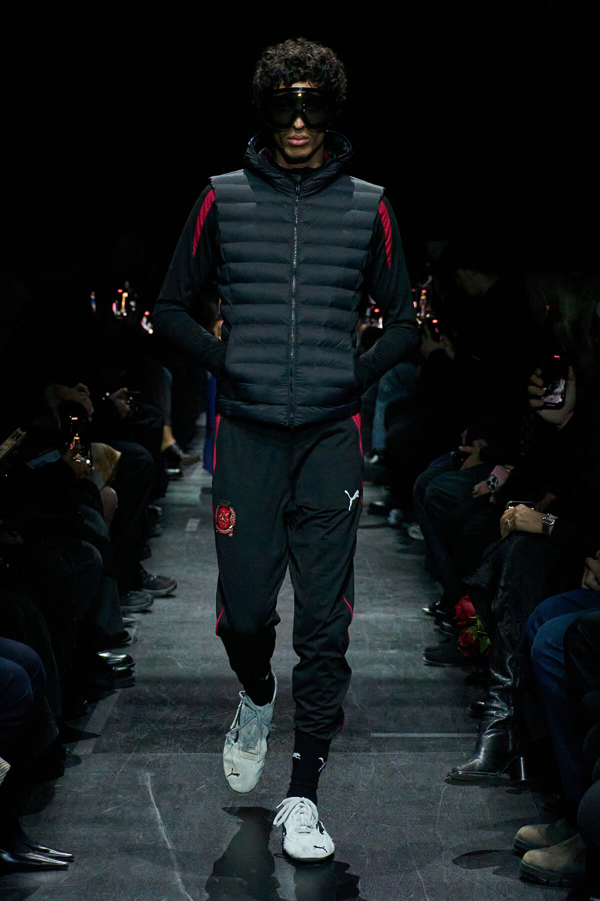 Male model walks the runway at night in a black padded vest over a black tracksuit with red accents and white sneakers.
