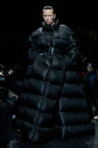 Model on a runway wearing an oversized black puffer coat with a high collar and a wide belt; dark background.