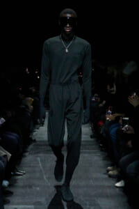 Male model walking a fashion show runway in an all-black outfit with oversized visor sunglasses and a silver pendant necklace.