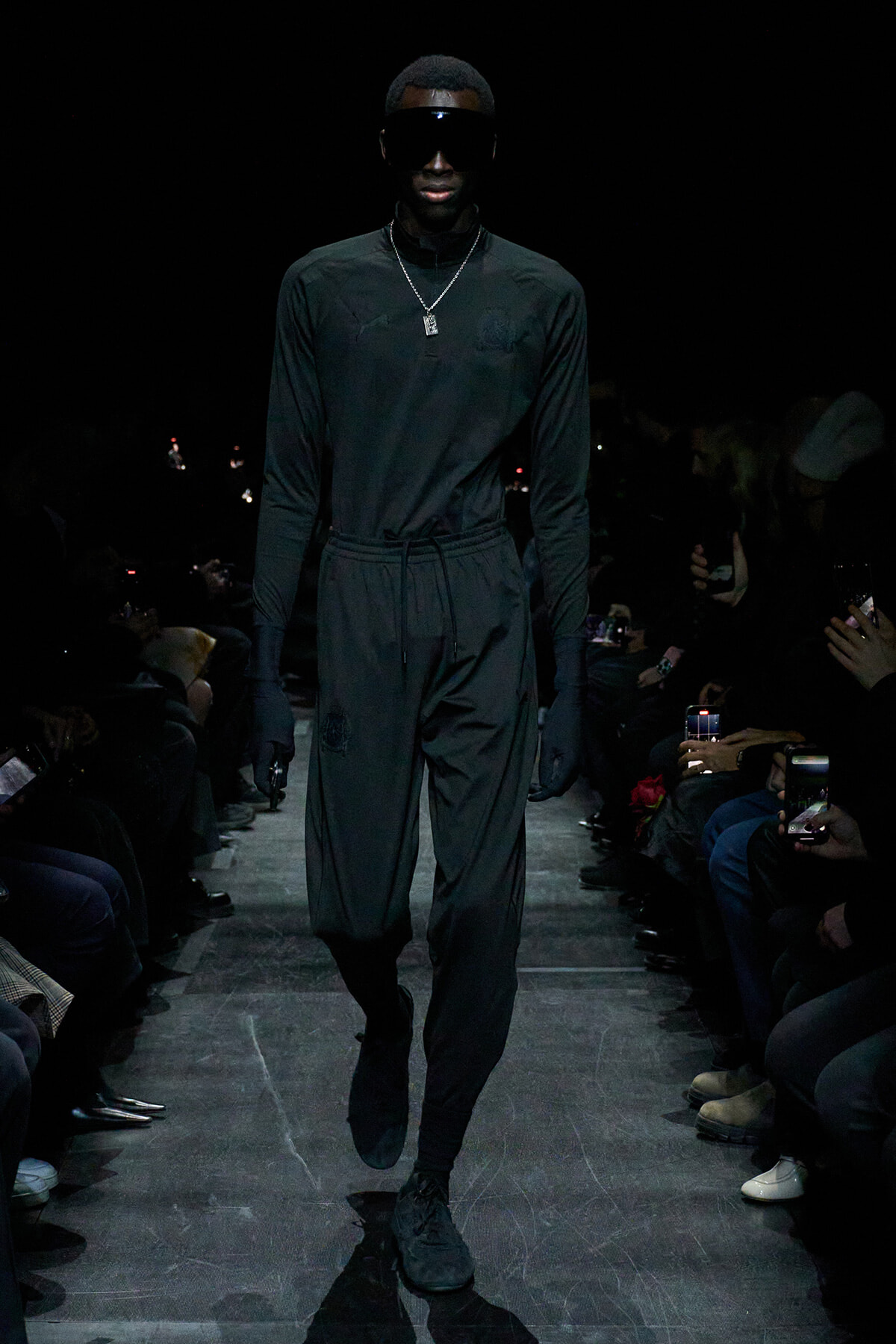 Male model walking a fashion show runway in an all-black outfit with oversized visor sunglasses and a silver pendant necklace.