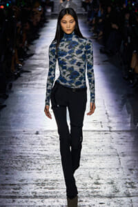 Model walks a fashion runway wearing a blue floral turtleneck top with black pants and subtle leather details at the hips.