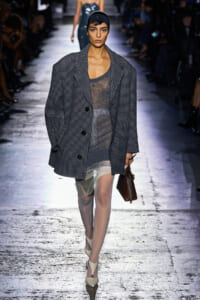Model walks the runway in a gray oversized checkered coat over a sheer gray dress, headband, and beige heels, carrying a brown handbag.