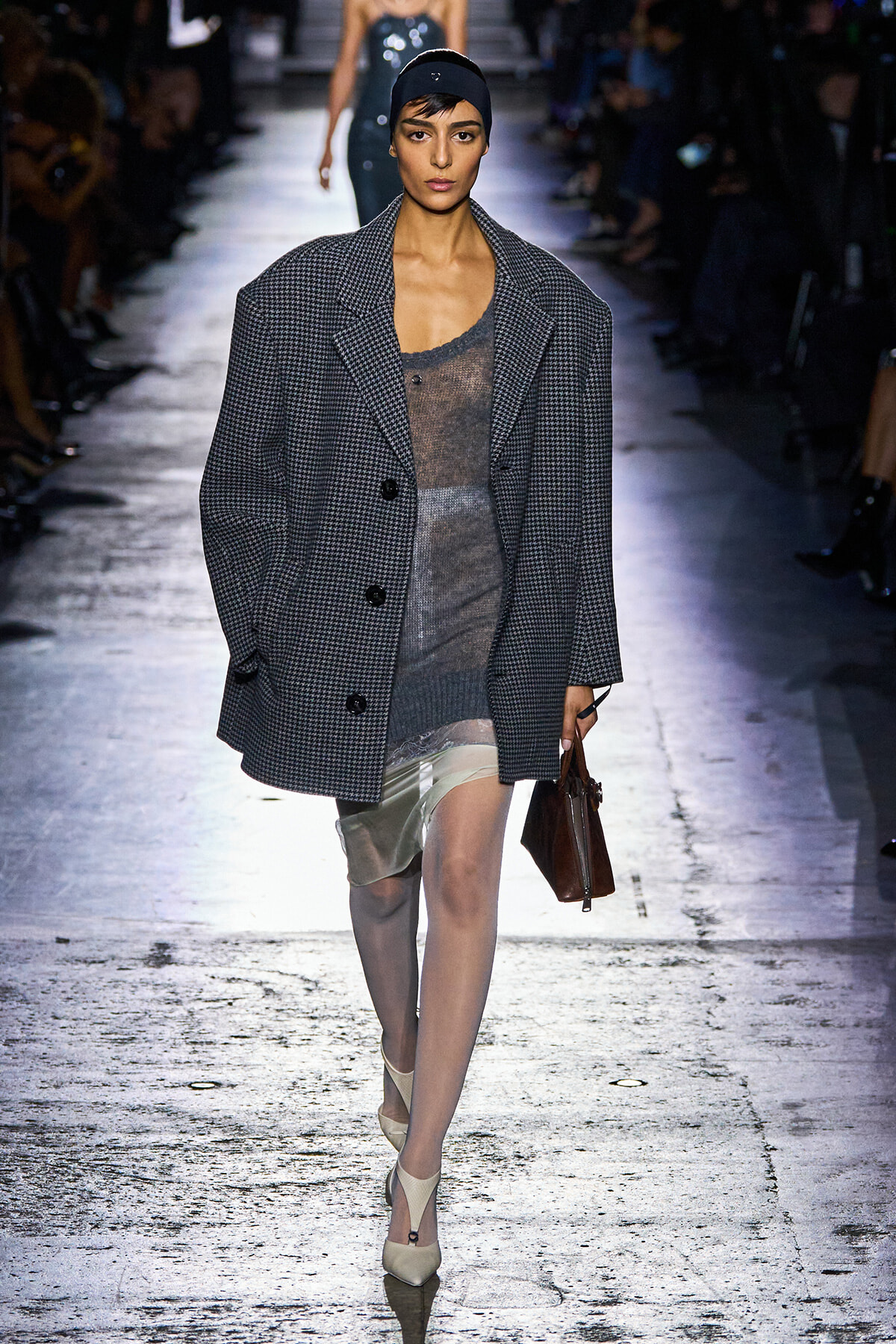 Model walks the runway in a gray oversized checkered coat over a sheer gray dress, headband, and beige heels, carrying a brown handbag.