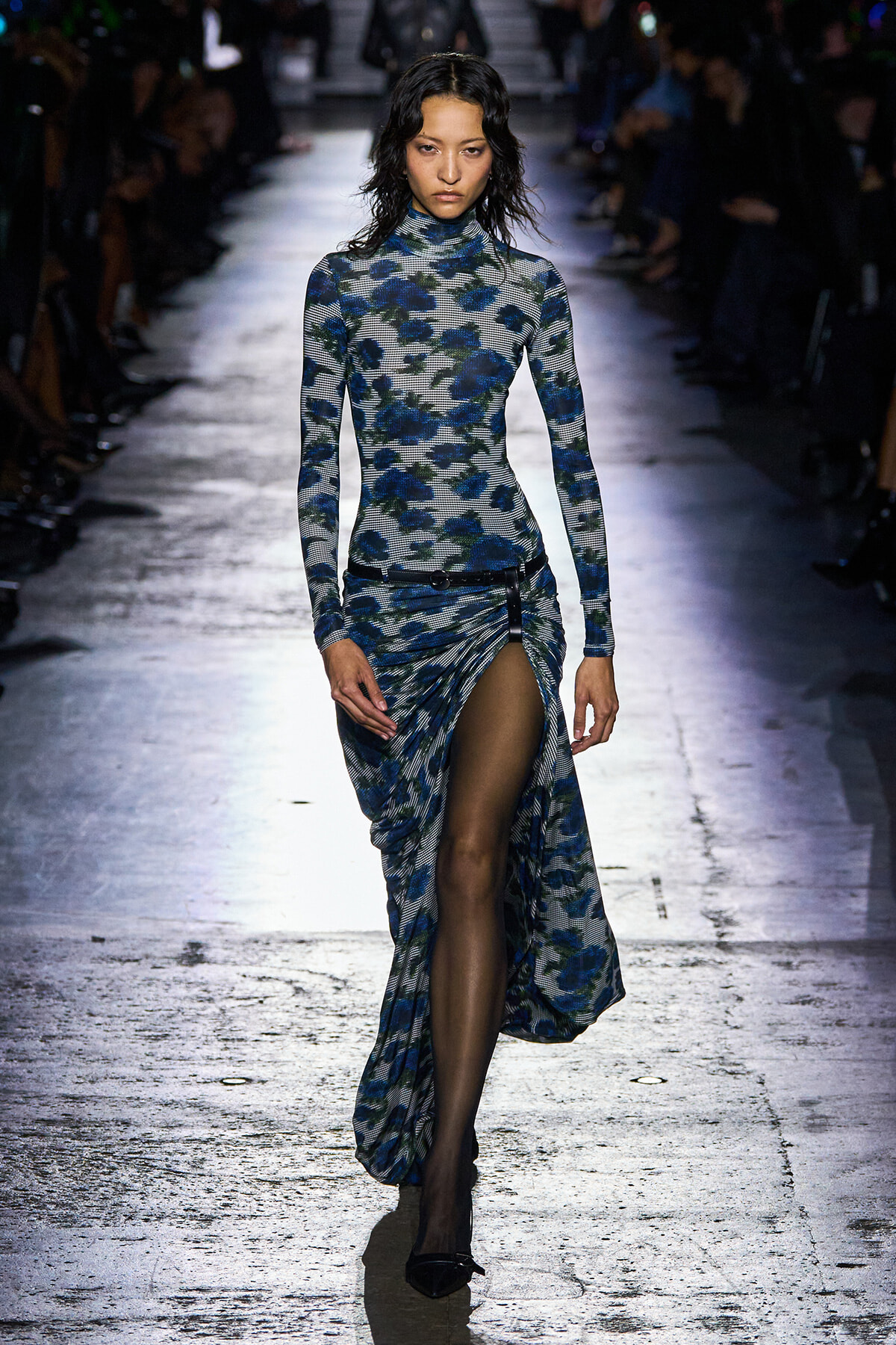Model walking a runway in a blue floral long-sleeve turtleneck dress with a high slit, black belt, and black heels.