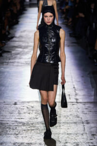 Model on a runway wearing a glossy black sleeveless hooded vest, paired with a short pinstriped skirt and black accessories.