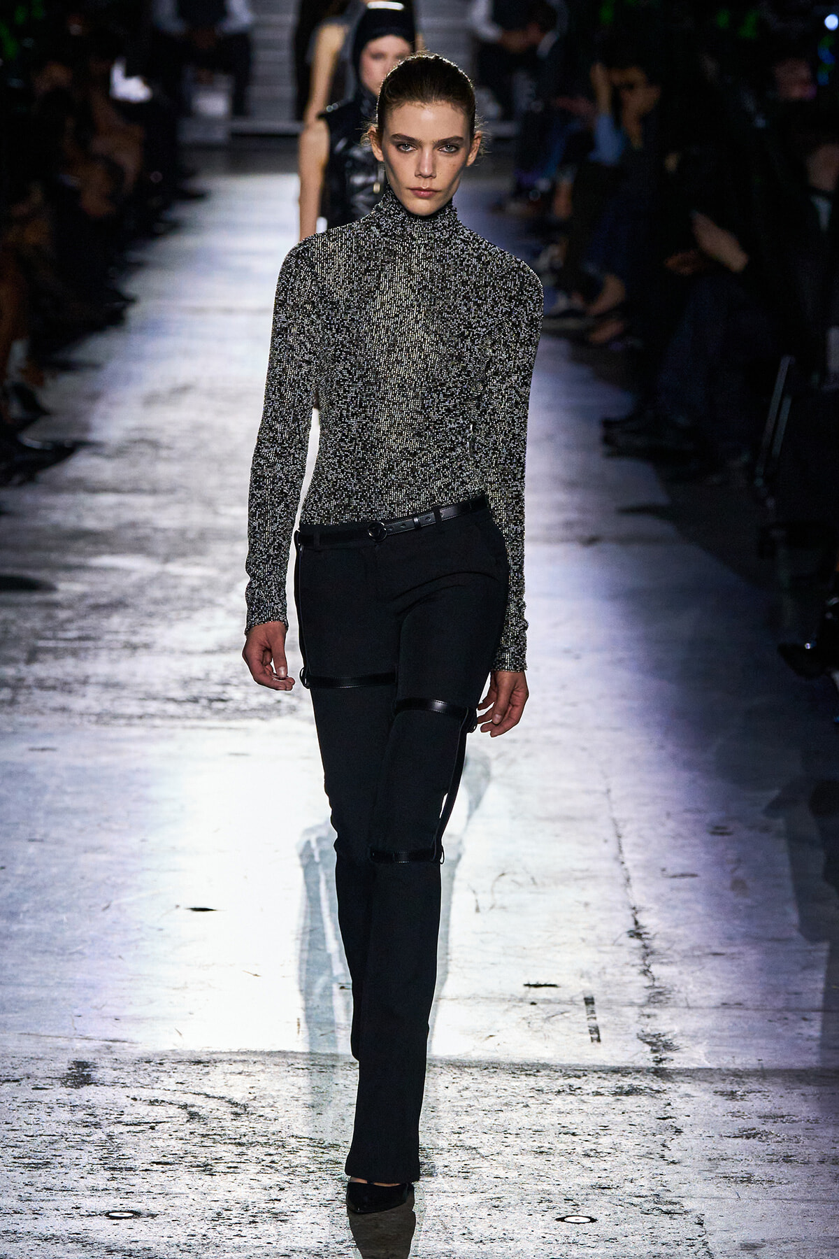 Model walks a runway in a glittery silver-black turtleneck sweater and black tailored pants with strap accents.