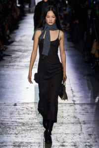 Model walking a fashion runway in a dark pinstripe sleeveless dress with a gray scarf around the neck and black boots, carrying a small bag and tote.