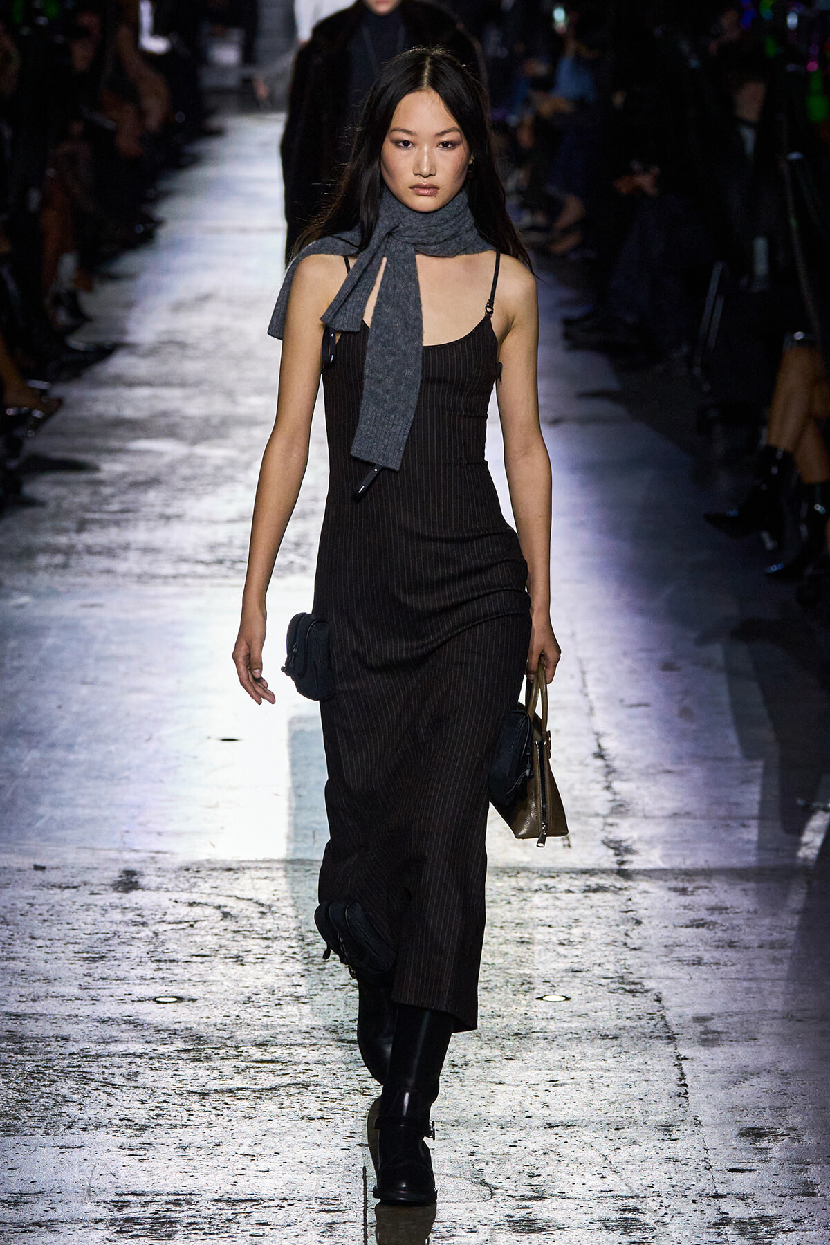 Model walking a fashion runway in a dark pinstripe sleeveless dress with a gray scarf around the neck and black boots, carrying a small bag and tote.