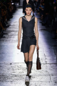 Runway model walking in a dark sleeveless top with sailor collar, a pleated wrap skirt, headband, and a brown handbag.