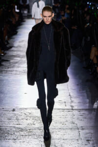 Model walking a fashion show runway in a long black faux-fur coat over a black turtleneck and pants, accessorized with a long silver pendant necklace.