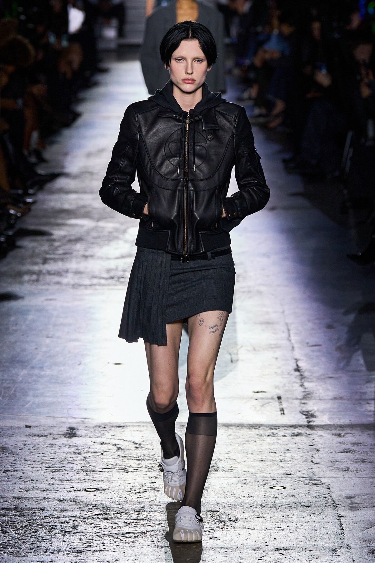 Model walks a fashion runway wearing a black leather bomber-style jacket and a dark gray pleated asymmetrical mini skirt, paired with mismatched legwear and white sneakers.