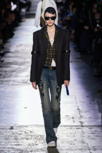 Runway model wearing a dark pinstriped blazer over a yellow plaid shirt, embroidered jeans, white sneakers, black cap and oversized sunglasses.