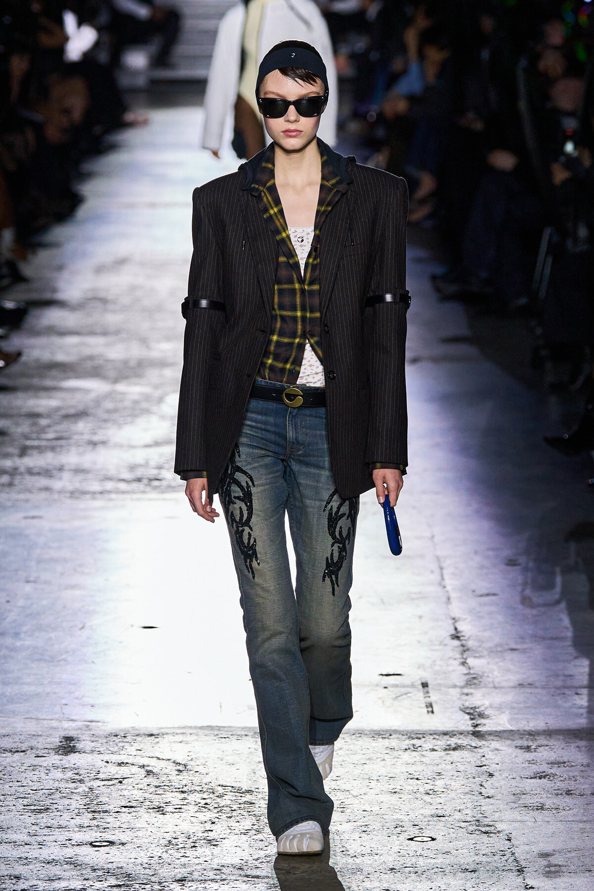 Runway model wearing a dark pinstriped blazer over a yellow plaid shirt, embroidered jeans, white sneakers, black cap and oversized sunglasses.