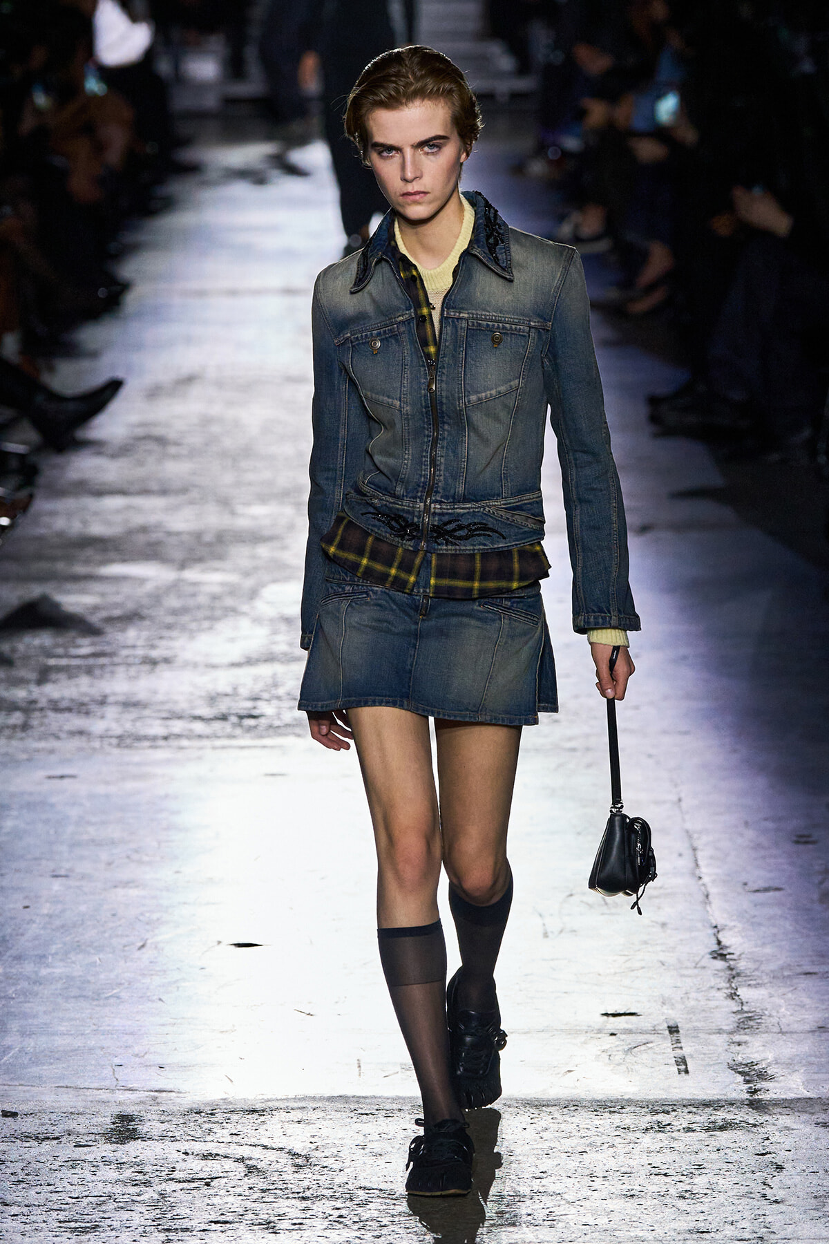 Model walking a fashion runway in a blue denim jacket and matching mini skirt, with a plaid underlayer and a small black bag in hand.