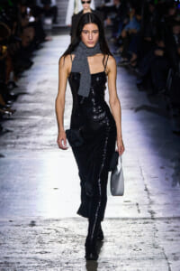 Model walking runway in a black sequined slip dress, gray scarf, black boots, and small gray handbag.