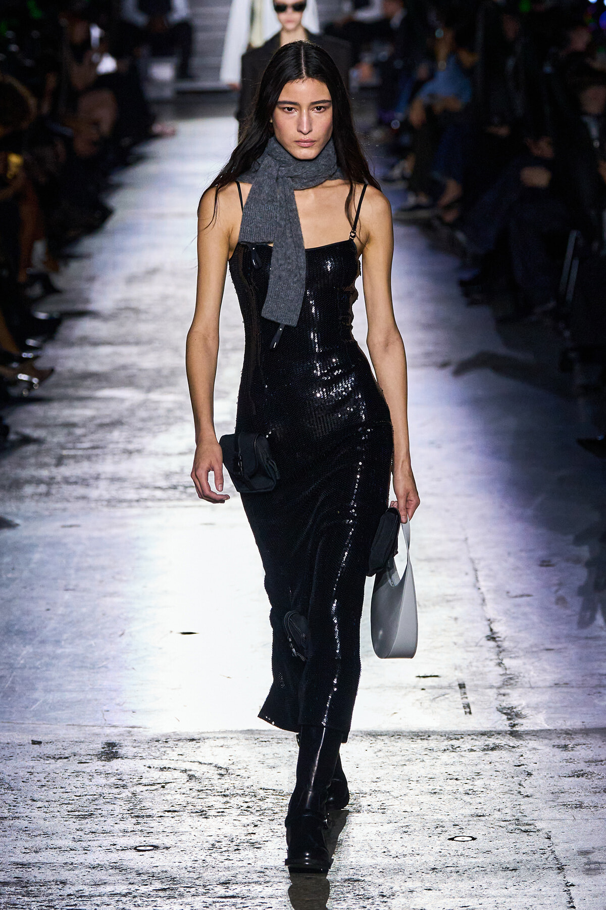 Model walking runway in a black sequined slip dress, gray scarf, black boots, and small gray handbag.