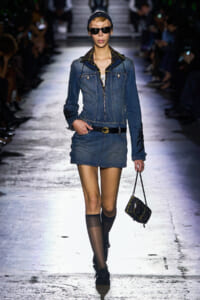 Model walking a fashion runway in a denim jacket and matching mini skirt, black belt, sunglasses, and a plaid bag accessory on a glossy catwalk.
