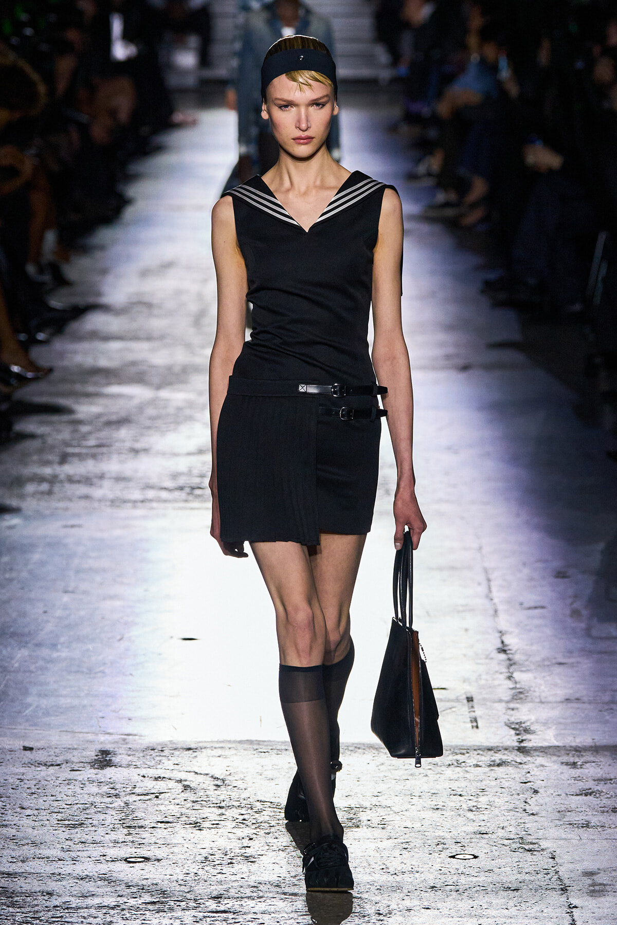 Model walking the runway in a black sleeveless top with a striped sailor collar and a black mini skirt, accessorized with a belt and sheer knee-high socks, carrying a black handbag.