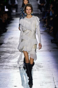 Model walking down a runway in a white, layered ruffled dress with pleats and long sleeves, paired with black knee-high boots.