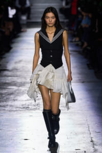 Model walking runway in black sleeveless vest with sailor collar and a white tiered ruffled skirt, paired with knee-high black boots and a light gray bag.