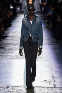 Model walks a fashion runway wearing a blue denim jacket over a white top, dark pinstripe pants, black sunglasses, and a black headband with a hoodie detail at the jacket cuffs