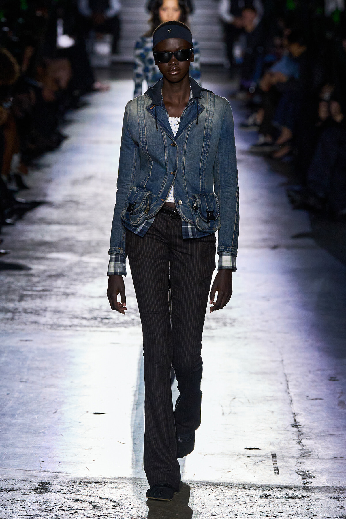 Model walks a fashion runway wearing a blue denim jacket over a white top, dark pinstripe pants, black sunglasses, and a black headband with a hoodie detail at the jacket cuffs