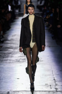 Model walking a fashion runway in an oversized pinstripe blazer over a beige bodysuit, with a plaid shirt and sheer tights, plus green leg pouches and black heels.