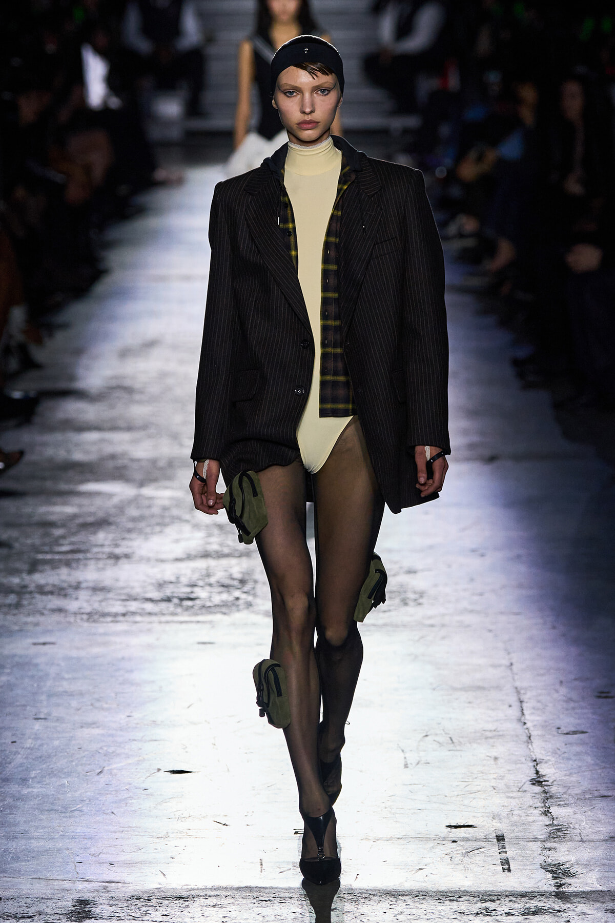 Model walking a fashion runway in an oversized pinstripe blazer over a beige bodysuit, with a plaid shirt and sheer tights, plus green leg pouches and black heels.
