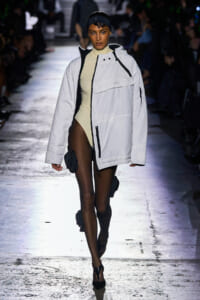 Model walks a runway wearing an oversized white jacket over a beige bodysuit, black headband, and leg pouches.