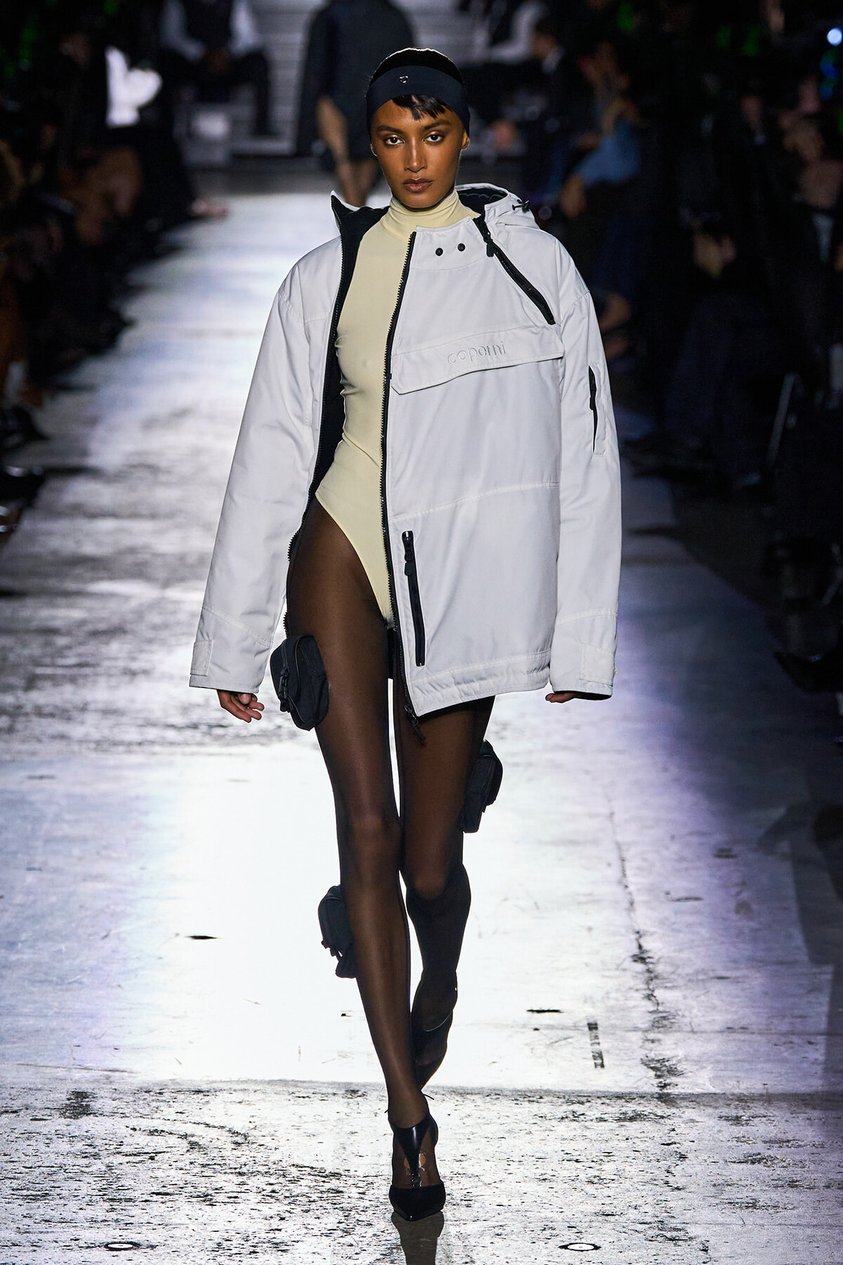 Model walks a runway wearing an oversized white jacket over a beige bodysuit, black headband, and leg pouches.